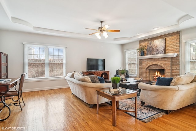 living room with a brick fireplace, light wood finished floors, ceiling fan, a healthy amount of sunlight, and a tray ceiling