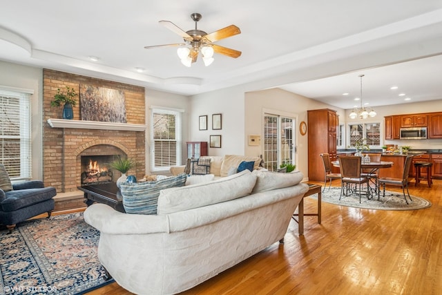 living room with recessed lighting, light wood-type flooring, ceiling fan with notable chandelier, a raised ceiling, and a fireplace