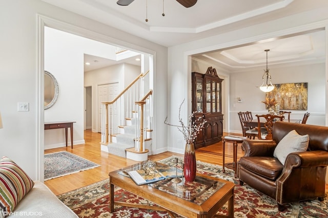 living room with stairway, a raised ceiling, light wood-type flooring, baseboards, and ceiling fan