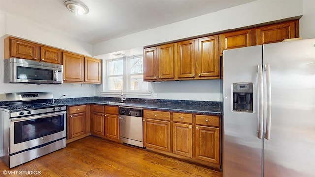 kitchen featuring brown cabinets, appliances with stainless steel finishes, a sink, dark stone counters, and dark wood-style floors