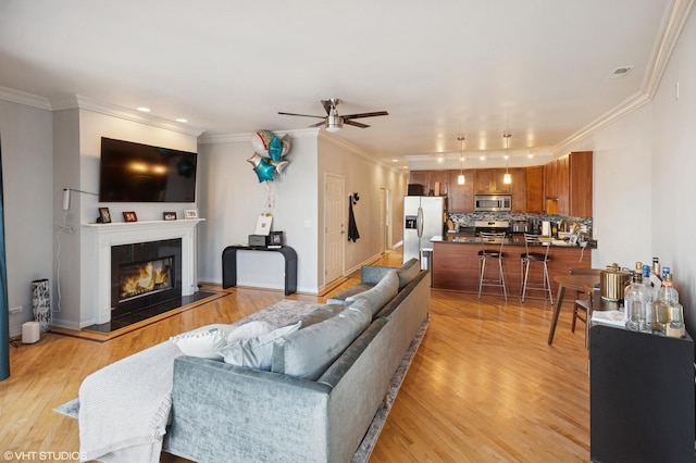 living area featuring crown molding, ceiling fan, light wood-type flooring, a tiled fireplace, and baseboards