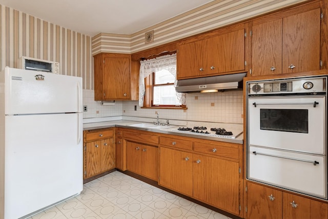 kitchen featuring a sink, a warming drawer, under cabinet range hood, white appliances, and wallpapered walls