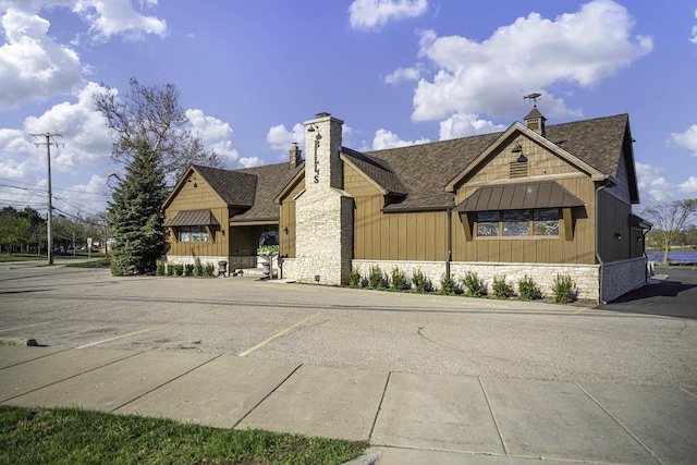 view of front of property with a shingled roof, stone siding, uncovered parking, a chimney, and board and batten siding