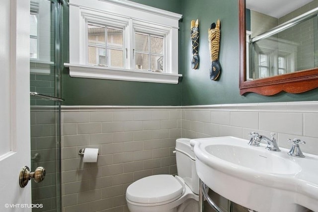 bathroom with a shower with door, toilet, wainscoting, tile walls, and a sink