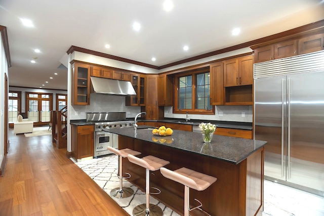 1745 W Cornelia Ave, Chicago, IL 60657 - kitchen featuring high end appliances, under cabinet range hood, brown cabinetry, decorative backsplash, and a center island with sink