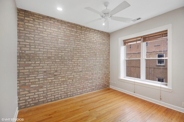 spare room with wood-type flooring, baseboards, brick wall, visible vents, and a ceiling fan