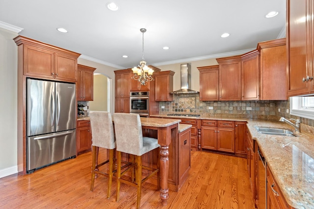 kitchen with wall chimney range hood, brown cabinets, appliances with stainless steel finishes, an inviting chandelier, and a sink