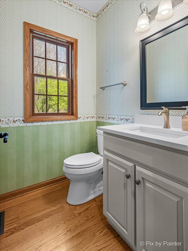 half bathroom with wainscoting, wallpapered walls, wood finished floors, and toilet