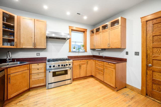 kitchen featuring high end range, wall chimney exhaust hood, dark countertops, a sink, and light wood finished floors