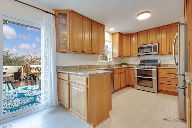 kitchen featuring glass insert cabinets, a textured ceiling, light stone countertops, stainless steel appliances, and a sink