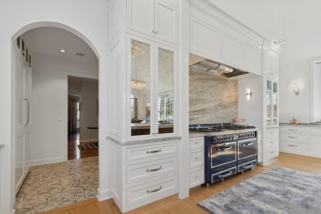 kitchen featuring range with two ovens, light wood-type flooring, backsplash, arched walkways, and white cabinets