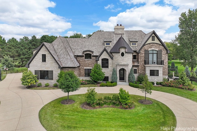 french country inspired facade with curved driveway, stone siding, a front lawn, and a chimney