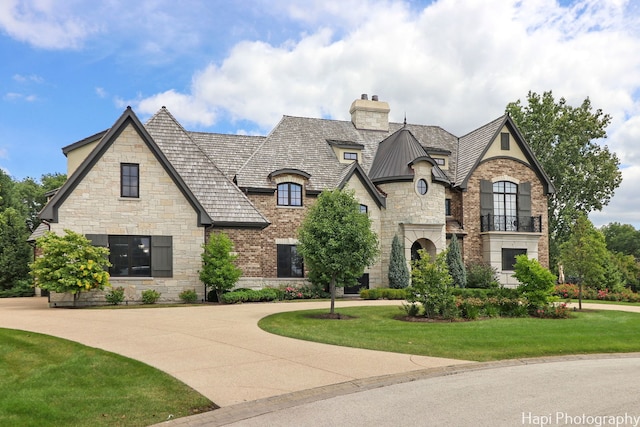french country inspired facade with a balcony, a chimney, curved driveway, stone siding, and a front yard