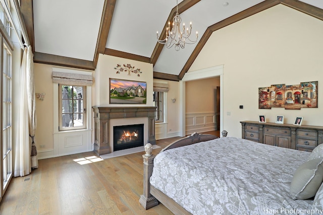 bedroom with an inviting chandelier, a decorative wall, light wood-type flooring, beamed ceiling, and a glass covered fireplace