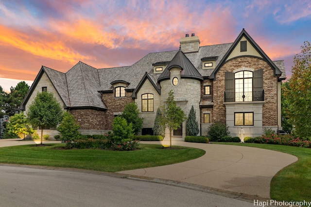 french country home featuring a yard, stone siding, a chimney, a balcony, and curved driveway