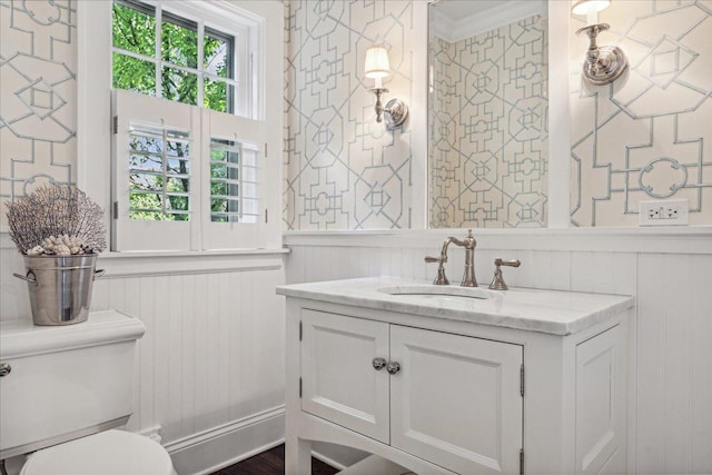 bathroom with toilet, vanity, wallpapered walls, and a wainscoted wall