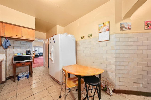 kitchen with light tile patterned floors, tile walls, stainless steel microwave, white refrigerator with ice dispenser, and a wainscoted wall