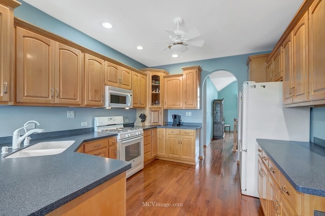 kitchen with arched walkways, a sink, dark countertops, white appliances, and wood finished floors