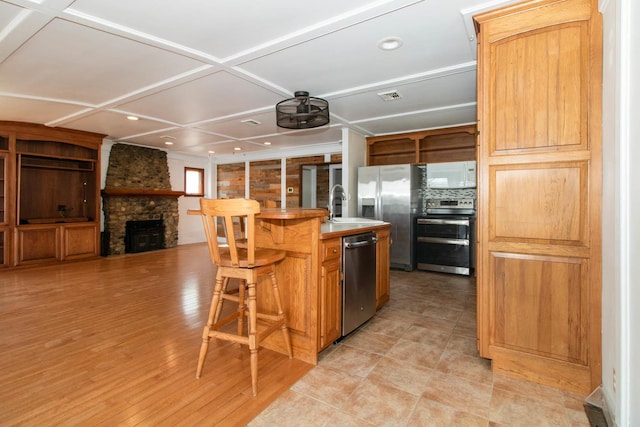 kitchen featuring a center island with sink, appliances with stainless steel finishes, open shelves, a stone fireplace, and a breakfast bar area