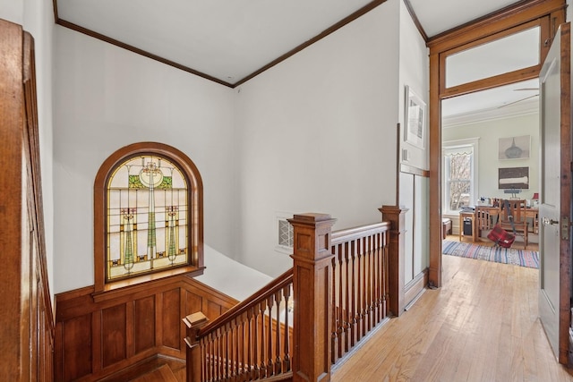 hallway featuring ornamental molding, light wood-style flooring, an upstairs landing, and a wainscoted wall