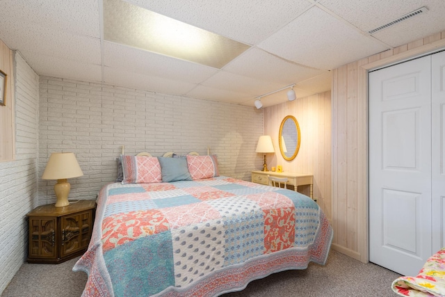 carpeted bedroom with a drop ceiling, track lighting, brick wall, and wood walls
