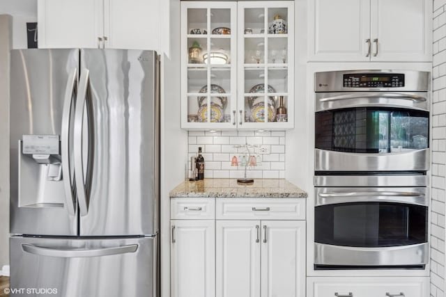 kitchen featuring glass insert cabinets, tasteful backsplash, white cabinetry, appliances with stainless steel finishes, and light stone counters