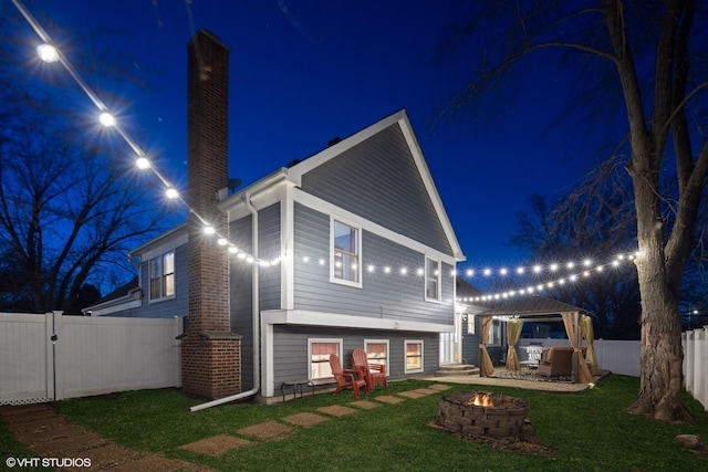 back of house at night featuring a yard, a fenced backyard, an outdoor fire pit, a gazebo, and a chimney