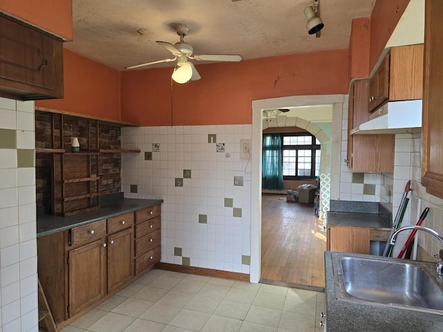 kitchen featuring brown cabinetry, a sink, a ceiling fan, and tile walls