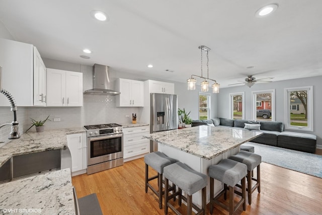 kitchen featuring wall chimney range hood, open floor plan, light wood-style floors, a breakfast bar area, and appliances with stainless steel finishes
