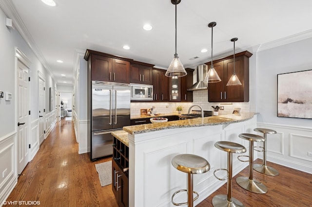 kitchen with a decorative wall, wall chimney exhaust hood, dark brown cabinets, a sink, and built in appliances