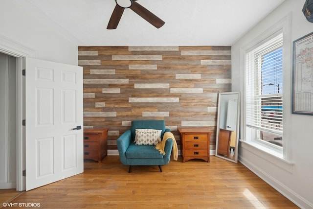 living area with light wood-style floors, an accent wall, a ceiling fan, wood walls, and baseboards