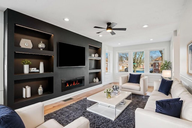 living area featuring a ceiling fan, visible vents, a glass covered fireplace, wood finished floors, and built in shelves