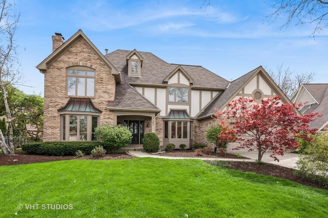 tudor-style house with a chimney, a front yard, brick siding, a shingled roof, and stucco siding