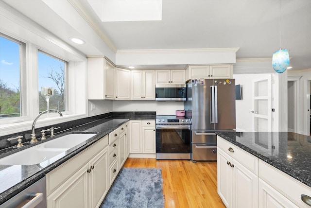 kitchen with a sink, stainless steel appliances, dark stone counters, ornamental molding, and light wood-type flooring