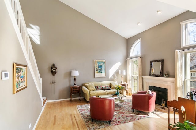 living area with baseboards, a tiled fireplace, stairway, light wood finished floors, and high vaulted ceiling