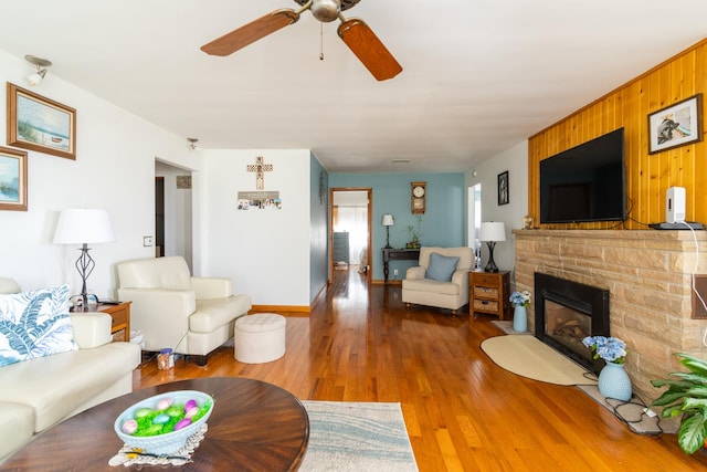 living area featuring baseboards, a stone fireplace, a ceiling fan, wood walls, and wood finished floors