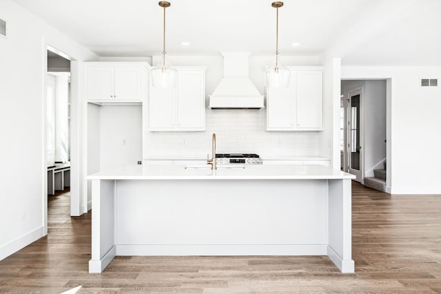 kitchen featuring visible vents, a center island with sink, light countertops, premium range hood, and decorative backsplash
