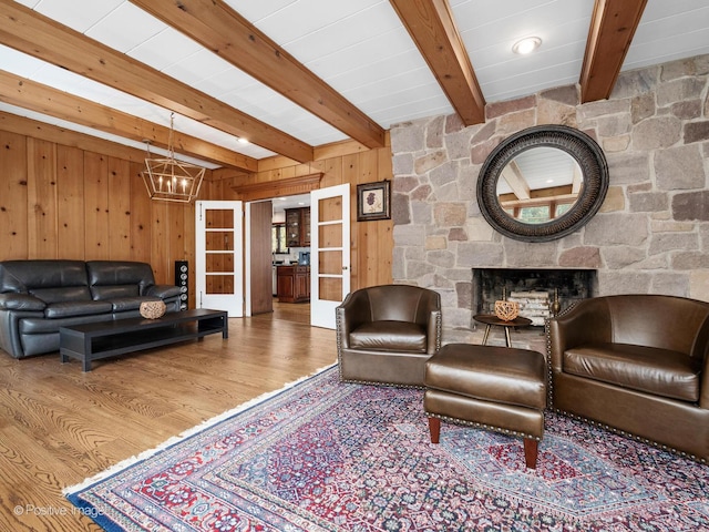 living area with a stone fireplace, wood walls, beam ceiling, wood finished floors, and a notable chandelier