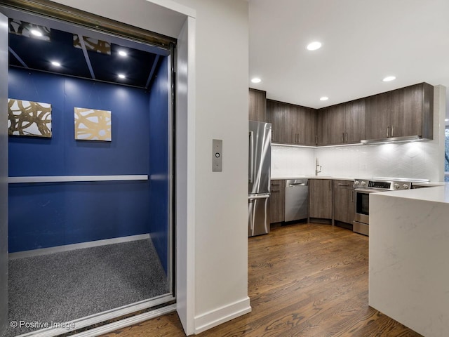 kitchen with elevator, decorative backsplash, stainless steel appliances, light countertops, and dark wood-style flooring