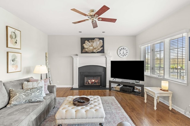 living area featuring a fireplace with flush hearth, baseboards, a ceiling fan, and wood finished floors