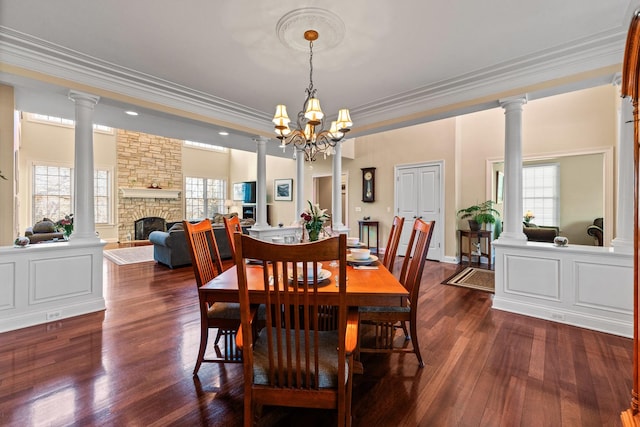 dining space with ornamental molding, a stone fireplace, decorative columns, and dark wood finished floors