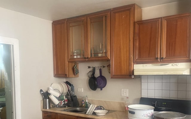 kitchen with glass insert cabinets, light countertops, under cabinet range hood, brown cabinetry, and stove