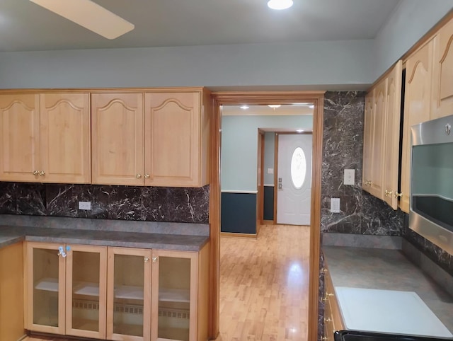kitchen featuring glass insert cabinets, light brown cabinets, dark countertops, stainless steel microwave, and light wood-style flooring