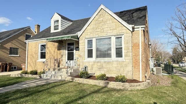 cape cod home with a front yard, central AC, stone siding, and a shingled roof