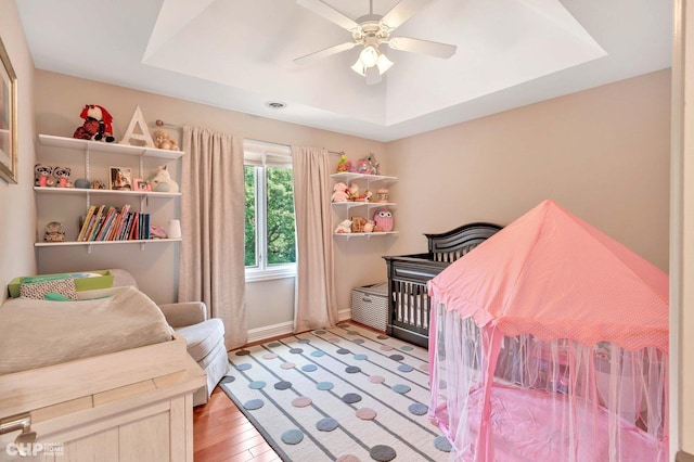 bedroom featuring baseboards, a raised ceiling, a ceiling fan, a crib, and light wood-style flooring