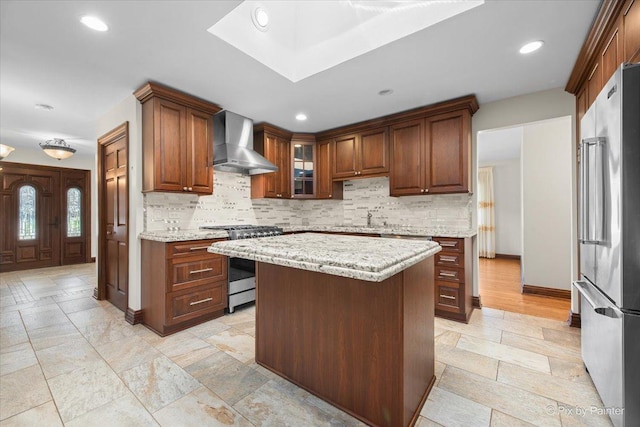 kitchen with wall chimney exhaust hood, glass insert cabinets, light stone countertops, stainless steel appliances, and backsplash