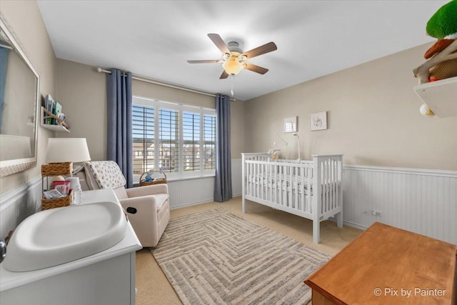bedroom featuring a nursery area, carpet flooring, ceiling fan, and a wainscoted wall