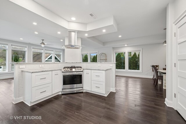 kitchen featuring wall chimney range hood, a ceiling fan, dark wood finished floors, stainless steel range with gas cooktop, and white cabinetry