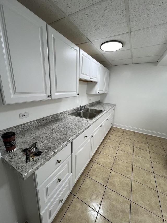 kitchen featuring baseboards, light tile patterned floors, white cabinets, a drop ceiling, and a sink