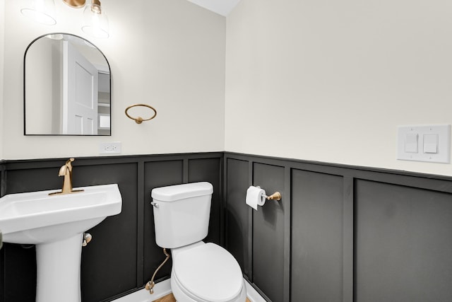 half bath with a decorative wall, toilet, a wainscoted wall, and a sink